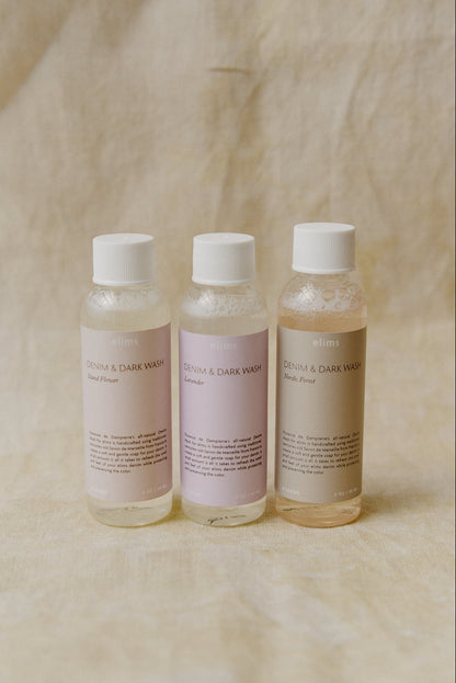 Three small clear bottles of Denim & Dark wash with white caps, arranged side by side on a beige fabric background.